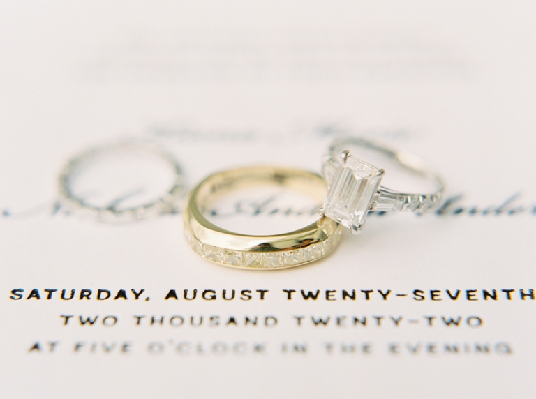 Emerald-cut diamond engagement ring and gold band on top of a wedding invitation